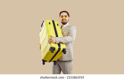 Handsome Young Businessman Going On Holiday Or Business Trip. Happy Attractive Man In Formal Suit Standing Isolated On Beige Background, Holding His Yellow Suitcase And Smiling. Travel Concept