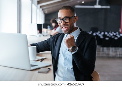 Handsome young businessman sitting at his ddesk, celebrating success with arms raised while looking at his laptop screen - Powered by Shutterstock