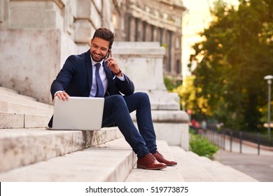 A handsome young businessman sitting next to his laptop on the stairs while talking on his phone