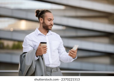 Handsome young businessman using smartphone and drinking coffee while walking outdoors during break, male entrepreneur browsing internet on mobile phone or answering business emails, copy space - Powered by Shutterstock