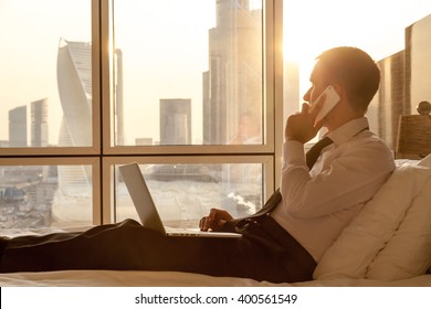 Handsome young businessman wearing formal white shirt and tie sitting on the bed with laptop in modern room. Self-employed person using smartphone in penthouse and looking at sunny city view in window - Powered by Shutterstock