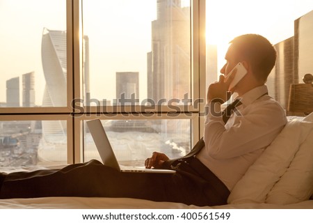 Handsome young businessman wearing formal white shirt and tie sitting on the bed with laptop in modern room. Self-employed person using smartphone in penthouse and looking at sunny city view in window