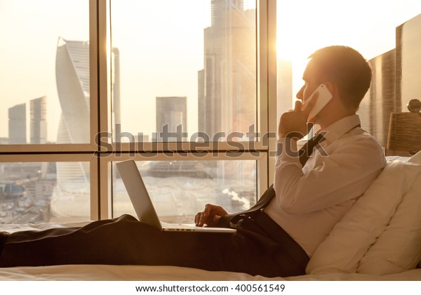 Handsome young businessman wearing formal white\
shirt and tie sitting on the bed with laptop in modern room.\
Self-employed person using smartphone in penthouse and looking at\
sunny city view in\
window