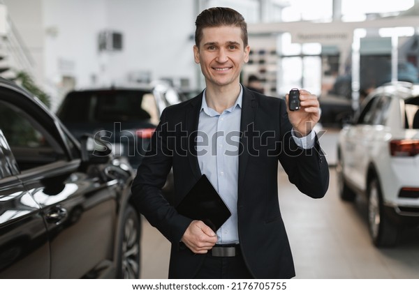 Handsome young car\
salesman showing car key at camera, selling autos at dealership.\
Portrait of amiling millennial manager presenting modern vehicles\
at showroom store