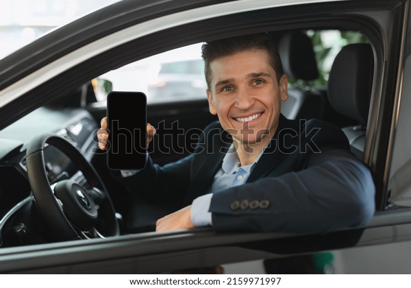Handsome young car\
salesman sitting in auto salon, showing blank mobile phone with\
mockup for design at modern dealership. Automobile insurance app or\
webite template