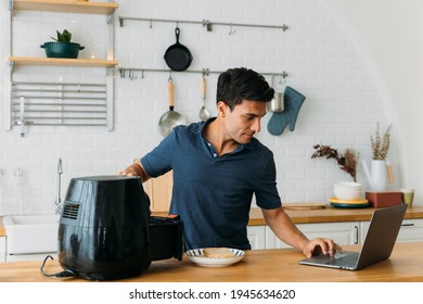 Handsome young caucasian man looking at instructions online on laptop on how to use modern air fryer to cook healthy food without using oil at home in kitchen