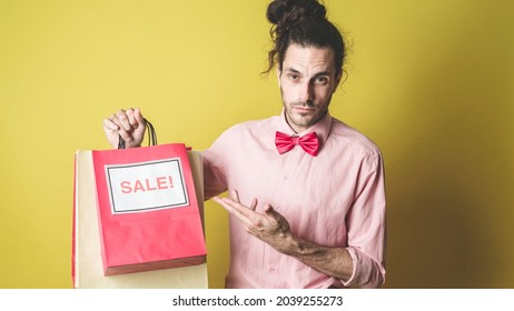 Handsome And Young Caucasian Personal Shopper Isolated Holding Multiple Gift Bags With The Sign Sale On Them. Wearing A Pink Shirt With A Red Bow Tie, Looking Straight At The Camera, Yellow Background