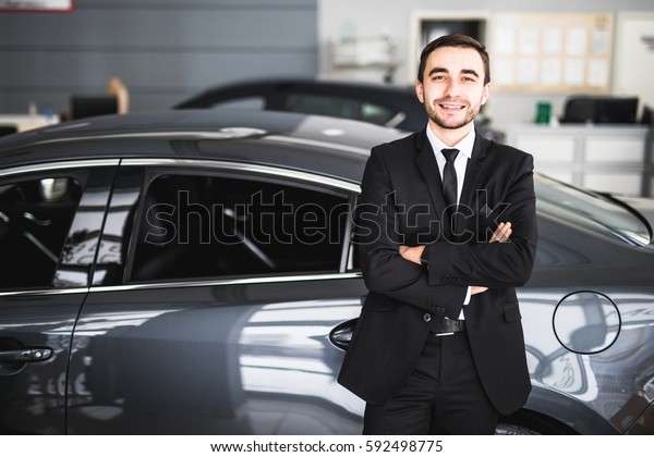 Handsome young classic car salesman standing at\
the dealership in front of new\
car
