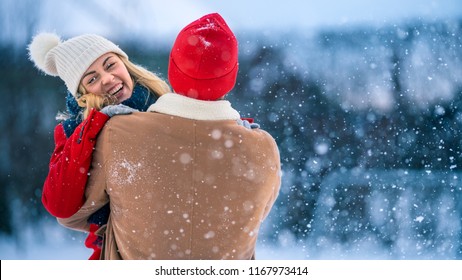 Handsome Young Couple Embrace And Happily Smile, Snow Falls On Them. Couple In Love On The Backyard Of Their Idyllic Home One Beautiful Winter Day.