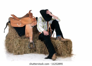 Handsome young cowboy with traditional outfit next to saddle sitting on hay.  Studio shot, white background.