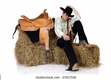 Handsome young cowboy with traditional outfit next to saddle sitting on hay.  Studio shot, white background.