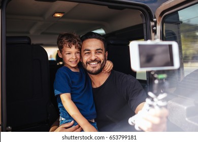 Handsome Young Dad And His Cute Little Son Sitting On Back Of The Car Making Selfie Using A Smart Phone. Father And Son On Road Trip Taking Selfie.