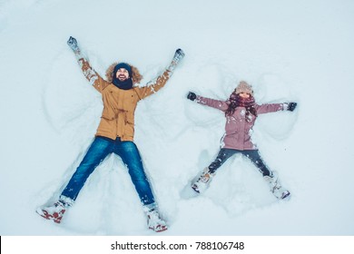 Handsome Young Dad And His Little Cute Daughter Are Having Fun Outdoor In Winter. Enjoying Spending Time Together While Making Snow Angels. Family Concept.