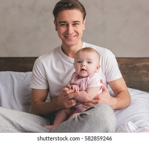 Handsome Young Dad Is Holding His Cute Baby In Arms, Looking At Camera And Smiling While Sitting On Bed