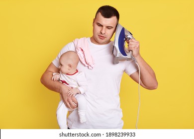 Handsome Young Dad Looking After Newborn Baby, Needs Ironing, Holds Iron Near Ear Like Mobile Phone, Imagines He Talks To Somebody,, Posing Isolated Over Yellow Background.