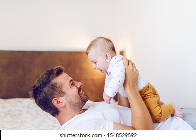 Handsome Young Dad Lying In Bed In Bedroom And Lifting His Loving Laughing Baby Boy Six Months Old. Unconditional Love Concept.