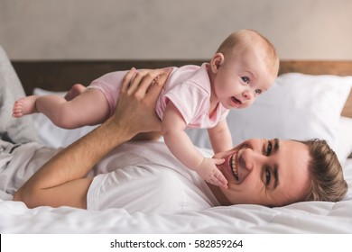 Handsome Young Dad Is Playing With His Cute Baby, Looking At Camera And Smiling