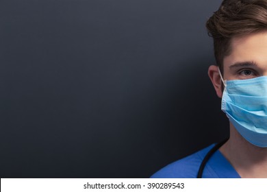Handsome young doctor in blue medical uniform and mask is looking at camera, standing against blackboard, cropped, close-up