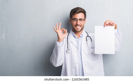 Handsome Young Doctor Man Over Grey Grunge Wall Holding Blank Sheet Contract Doing Ok Sign With Fingers, Excellent Symbol