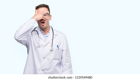 Handsome Young Doctor Man Peeking In Shock Covering Face And Eyes With Hand, Looking Through Fingers With Embarrassed Expression.