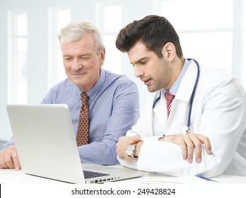 Handsome Young Doctor Sitting In Front Of Computer And Consulting With Old Patient.