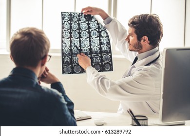Handsome young doctor in white coat is showing x-ray image to his patient while working in office