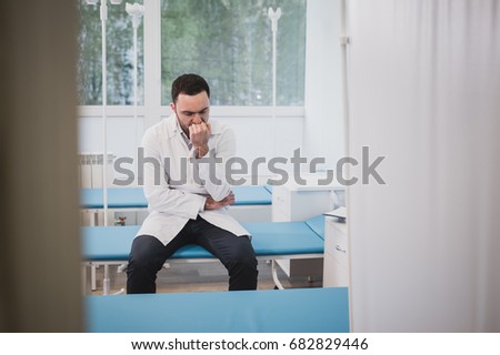 Handsome young doctor in white coat is sitting sadly in hospital ward