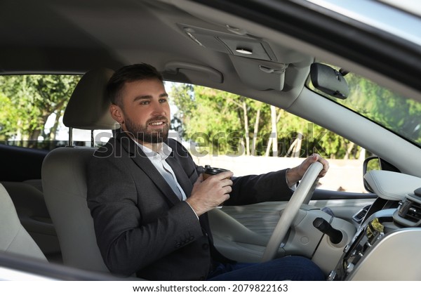 Handsome young driver with cup of drink sitting in\
modern car