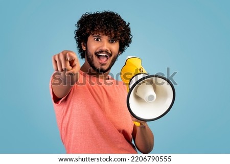 Handsome young eastern guy shouting through yellow loudspeaker and gesturing, pointing at camera, hindu man making announcement on blue studio background, copy space