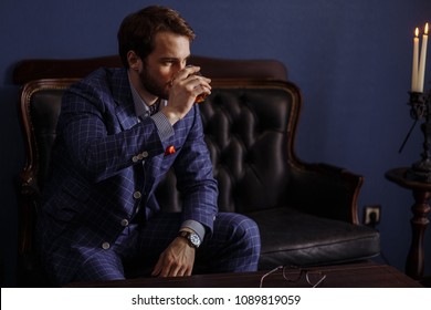 Handsome Young Elegant Man In A Classic Suit Drinking Beverage Sitting At Chesterfield Settee In Restaurant