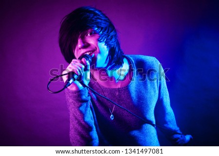 The handsome young emo guy is singing in microphone on purple and blue concert lighting.