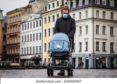 Handsome Young European Man In Winter Clothes On The Street With A Medical Face Mask On And Baby Carriage. 35-year-old Male In A Respirator To Protect Against Infection With  Coronavirus