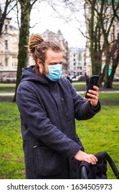 Handsome Young European Man In Winter Clothes On The Street With A Medical Face Mask On And Baby Carriage. 35-year-old Male In A Respirator To Protect Against Infection With  Coronavirus