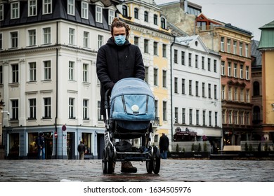 Handsome Young European Man In Winter Clothes On The Street With A Medical Face Mask On And Baby Carriage. 35-year-old Male In A Respirator To Protect Against Infection With  Coronavirus