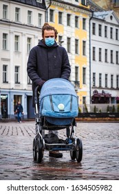 Handsome Young European Man In Winter Clothes On The Street With A Medical Face Mask On And Baby Carriage. 35-year-old Male In A Respirator To Protect Against Infection With  Coronavirus