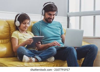 Handsome young father in casual clothes using a laptop and his cute little son using a tablet while sitting on a sofa in the room. Both in headphones.