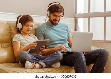 Handsome young father in casual clothes using a laptop and his cute little son using a tablet while sitting on a sofa in the room. Both in headphones.