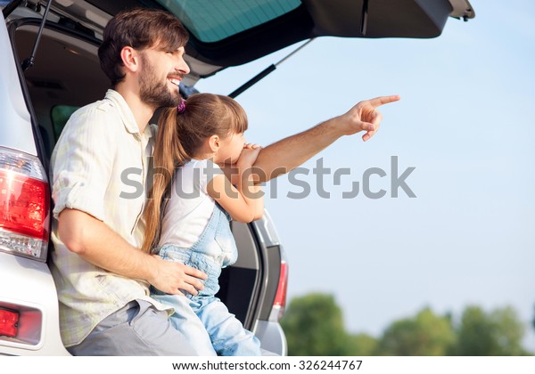 Handsome young father and his small daughter are\
sitting on open car boot in forest. The parent is pointing finger\
sideways and smiling. Girl is looking there with interest. Copy\
space in right side