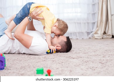 Handsome Young Father Is Playing With His Sun. He Is Lying On Flooring And Holding The Child Under Him With Joy. They Are Smiling. Copy Space In Right Side