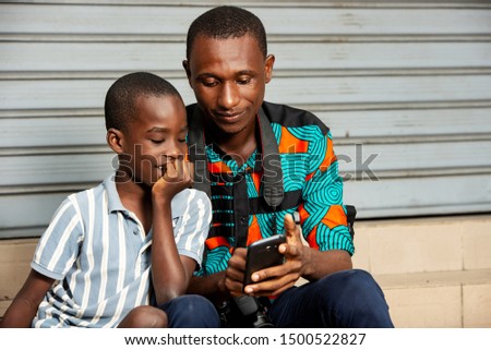 handsome young father sitting with his son, father showing images to his son on mobile phone in the street while smiling