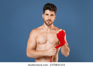 Handsome young fitness sporty strong guy bare-chested muscular sportsman boxer isolated on blue wall background studio. Workout sport motivation lifestyle concept. Reeling up sports bandages on hand - Powered by Shutterstock