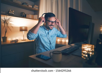 Handsome Young Freelancer Working Remotely At Night. He Is Putting On Headphones.