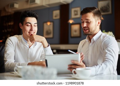 Handsome young friendly men in white shirts sitting at table in cafe and surfing tablet together