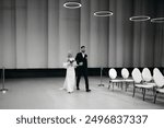 Handsome young groom in classic brown suit and pretty blond bride in vintage white dress with bouquet going to the wedding ceremony. Couple pre-wedding walking through ceremony hall with white chairs 