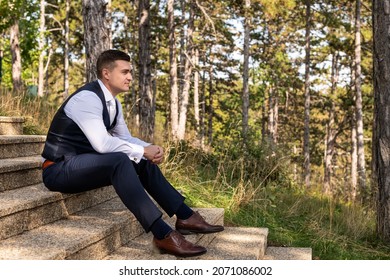 Handsome Young Groom Wearing Elegant And Stylish Dark Blue Suit Outdoors Portrait. Stylish Groom Sitting On Stairs Side View.