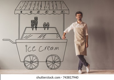 Handsome young guy in apron is leaning drawn ice cream cart  looking at camera   smiling while standing at the wall