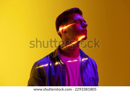 Handsome young guy in casual clothes with digital lights reflection on face standingover gradient yellow background. Energy. Concept of modern photography, art, cyberpunk, techno, creativity