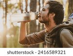 Handsome young guy drinking water while walking and traveling by pine forest with copy space. Rehydration with cold beverage during hiking backpacking alone in woods