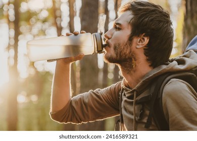 Handsome young guy drinking water while walking and traveling by pine forest with copy space. Rehydration with cold beverage during hiking backpacking alone in woods - Powered by Shutterstock