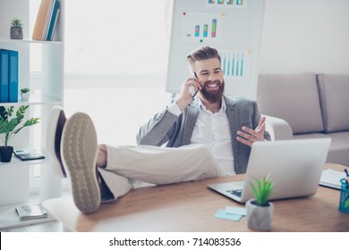 Handsome young guy in formalwear talking on mobile phone and telling about his achievement, sitting at the office with feet on the desk top, gesturing